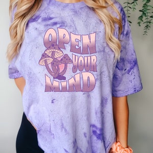 May include: A purple tie-dye t-shirt with the words "OPEN YOUR MIND" in a retro font. The shirt features a graphic of three mushrooms in shades of purple and pink. The shirt is a relaxed fit.