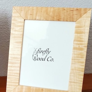 May include: A natural wood picture frame with a light wood finish. The frame has a simple design with a beveled edge. The frame is made by Firefly Wood Co.