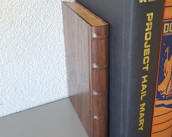 May include: A dark brown wooden bookend with a rounded spine and raised bands, positioned next to a black hardcover book titled Project Hail Mary by Andy Weir and an orange book with a graphic illustration on the spine.