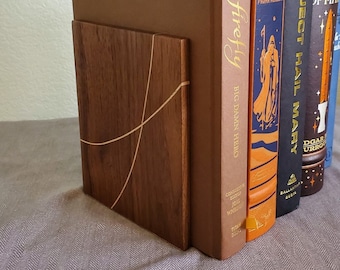 May include: A wooden bookend with a light-colored inlaid design supports a row of hardcover books. The books have various colors and titles, including "Firefly" and "Dune". The bookend is made of dark wood.