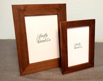 May include: Two wooden picture frames with a rich, reddish-brown finish. The frames have a simple, classic design and are of different sizes. The larger frame is positioned behind the smaller one. The text "firefly Wood Co." is printed in black on the white background.