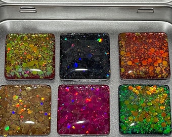 May include: A silver metal tin containing six square magnets with a glossy, multi-colored glitter finish in gold, black, orange, bronze, magenta, and green. The inside lid features a white label with the text M MAGNET MARKETPLACE.