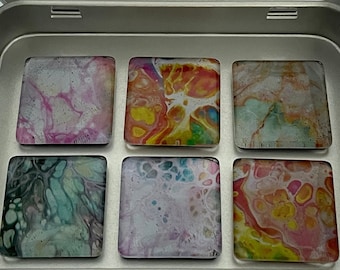 Può includere: Una scatola rettangolare in metallo argentato contenente sei magneti quadrati con disegni astratti realizzati con la tecnica del colaggio. Il coperchio interno presenta un'etichetta bianca con il testo MM MAGNET MARKETPLACE e il sito magnetmarketplace.etsy.com.