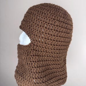 May include: A brown crocheted balaclava with a face opening. The balaclava is made with a simple stitch pattern.
