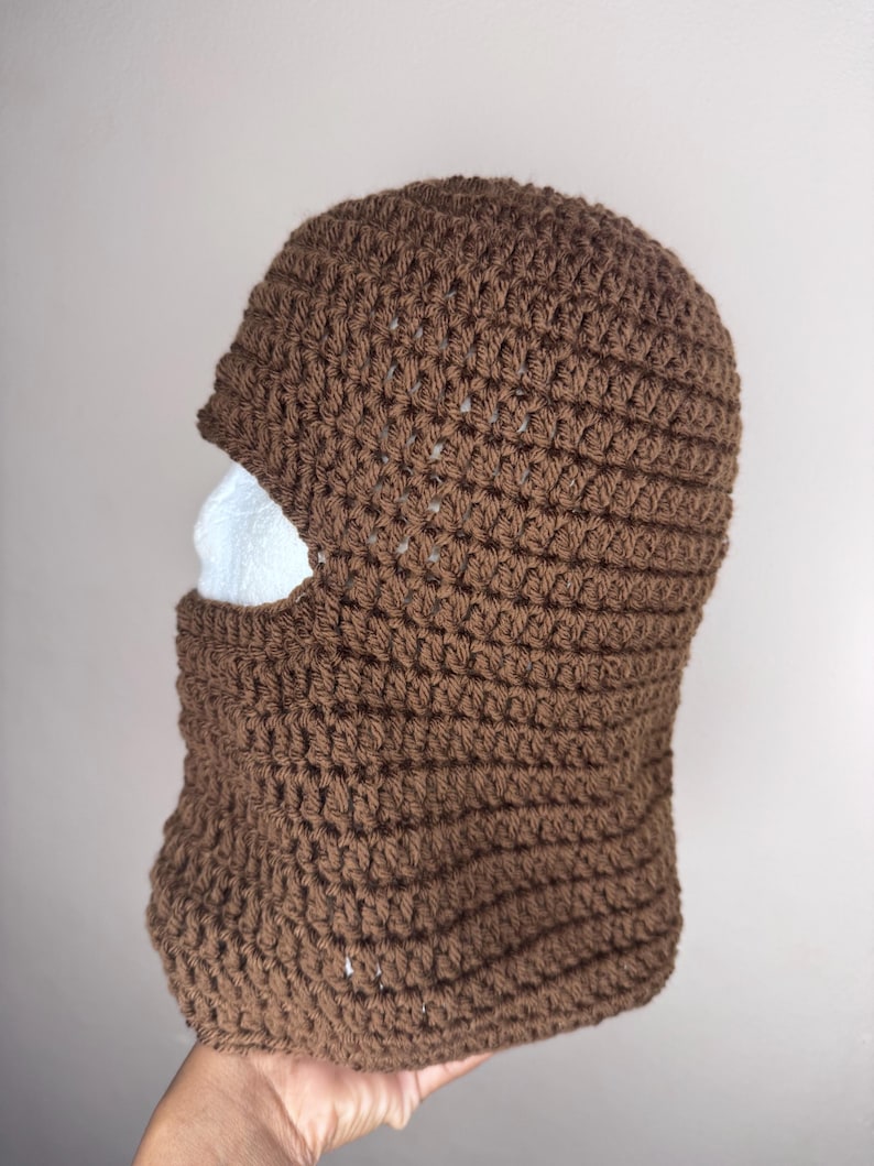 May include: A brown crocheted balaclava with a face opening. The balaclava is made with a simple stitch pattern.