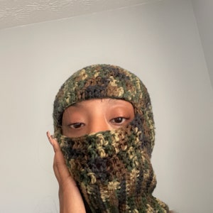 May include: A close-up of a person wearing a camouflage crochet balaclava. The balaclava is made of green, brown, and black yarn.