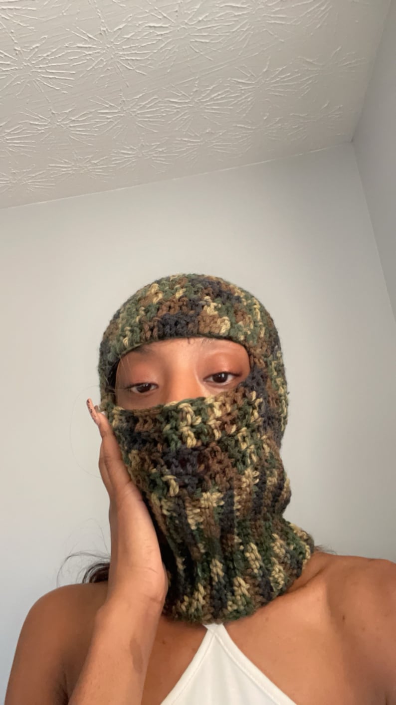 May include: A close-up of a person wearing a camouflage crochet balaclava. The balaclava is made of green, brown, and black yarn.