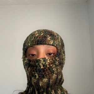 May include: A close-up of a person wearing a green, brown, and black camouflage crochet balaclava. The balaclava covers the person's face, leaving only their eyes visible. The person has long, dark nails with pink and white designs.