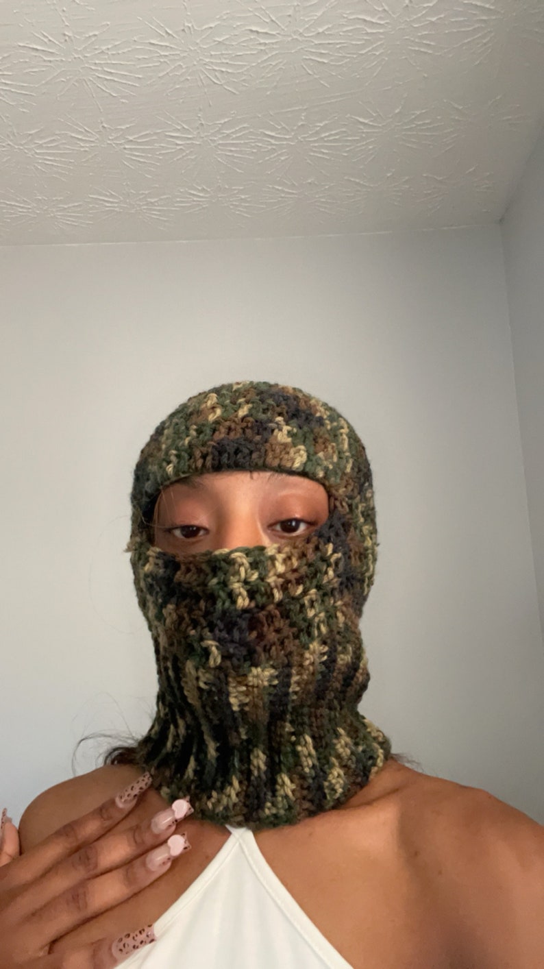 May include: A close-up of a person wearing a green, brown, and black camouflage crochet balaclava. The balaclava covers the person's face, leaving only their eyes visible. The person has long, dark nails with pink and white designs.