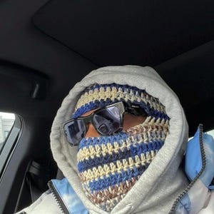 May include: A person wearing a gray hooded sweatshirt with a blue and brown striped crocheted face mask and black sunglasses. The person is sitting in a car.