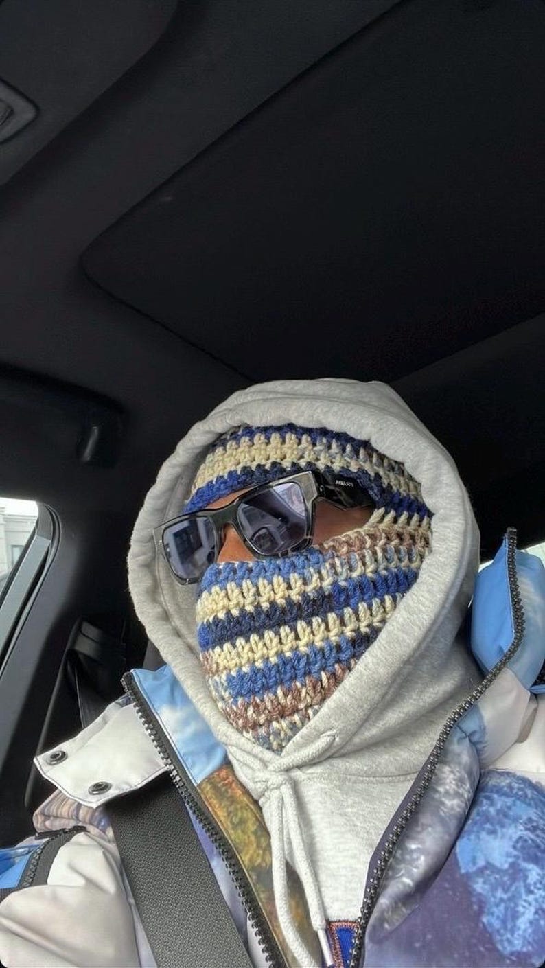 May include: A person wearing a gray hooded sweatshirt with a blue and brown striped crocheted face mask and black sunglasses. The person is sitting in a car.