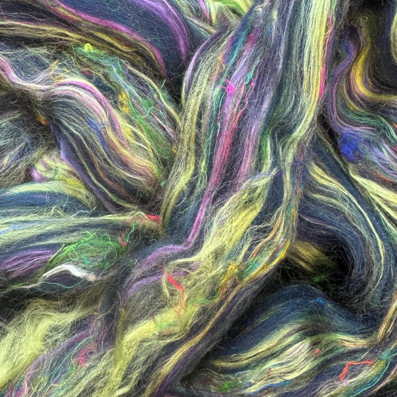 May include: A close-up of a colorful blend of yarn fibers in shades of black, green, yellow, and purple. The fibers are twisted and intertwined, creating a textured and dynamic visual.