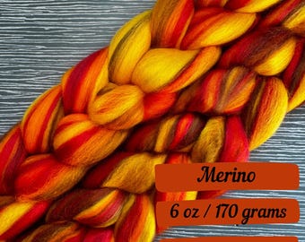 May include: A braided skein of Merino wool in vibrant autumn colors: yellow, orange, and red. The label indicates 6 oz / 170 grams of wool. The wool is soft and ideal for knitting, crocheting, or other fiber arts projects.