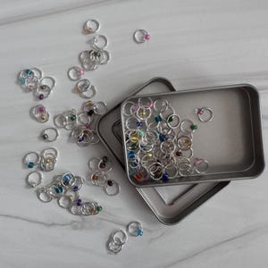 May include: A silver metal tin filled with colorful, beaded, stitch markers. The tin is open and some markers are scattered on a white surface.