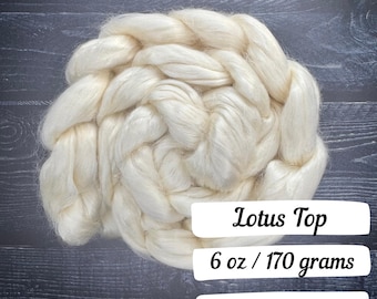 May include: Cream-colored Lotus Top fiber, weighing 6 oz. The fiber is arranged in a braided pattern. The text "Lotus Top" is displayed on a white rectangular label, along with the weight in ounces and grams.