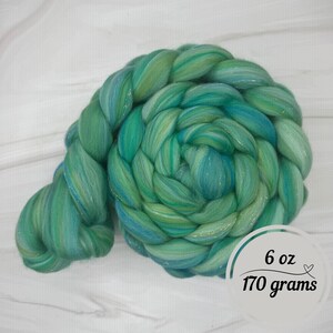 Pode incluir: Uma tran&ccedil;a vibrante de fibra tingida &agrave; m&atilde;o em tons de verde-azulado e verde. A fibra &eacute; um material macio e luxuoso, perfeito para fiar ou outras artes t&ecirc;xteis. A etiqueta indica um peso de 170 gramas (6 oz).