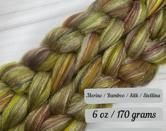 May include: Braided skein of yarn in shades of green, brown, and gold with a shimmering effect. The yarn is labeled as Merino, Bamboo, Silk, and Stellina, weighing 170 grams.