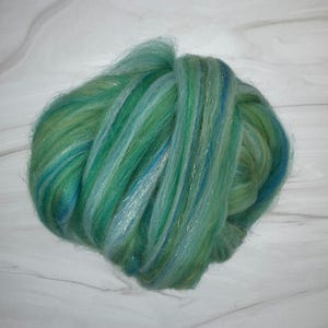 Pode incluir: Um feixe de fibra verde e azul, possivelmente l&atilde; ou material semelhante, &eacute; exibido sobre um fundo branco. As fibras s&atilde;o em camadas, criando uma apar&ecirc;ncia texturizada. As cores se misturam, com toques de ouro e prata.