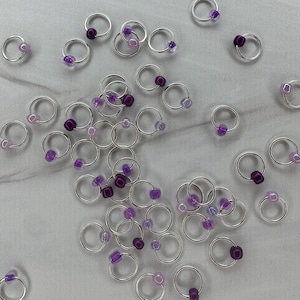 May include: A collection of small, silver-colored hoop earrings scattered on a light-colored surface. Each earring features a small, round, purple bead. Some beads are a darker shade of purple, while others are a lighter, more translucent hue.