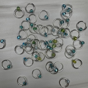May include: A collection of silver-colored hoop rings, each adorned with a small, colorful bead. The beads are in shades of blue, green, and yellow. The rings are scattered across a light gray surface, creating a visually appealing arrangement.