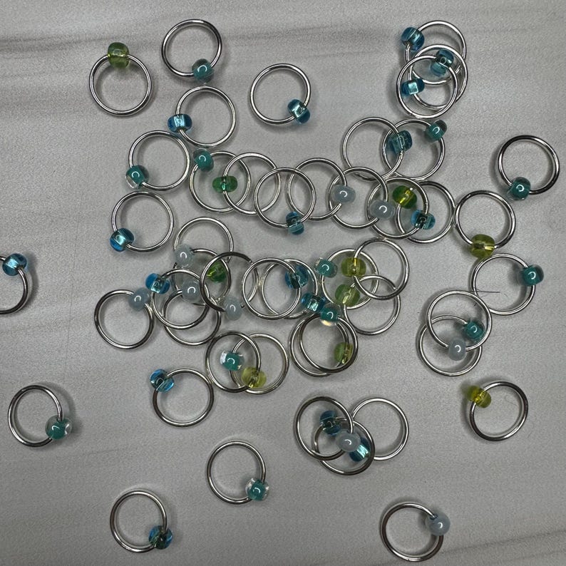 May include: A collection of silver-colored hoop rings, each adorned with a small, colorful bead. The beads are in shades of blue, green, and yellow. The rings are scattered across a light gray surface, creating a visually appealing arrangement.