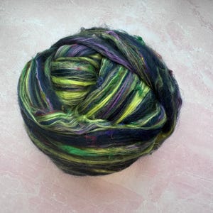 May include: A ball of colorful fiber with black, green, yellow, and purple strands. The fiber is arranged in a loose, fluffy ball.