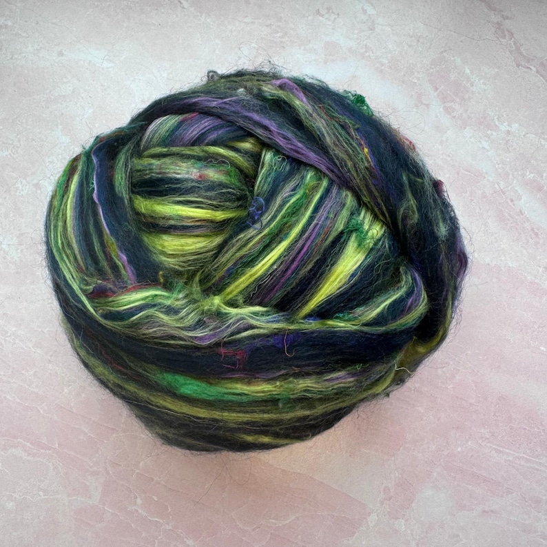 May include: A ball of colorful fiber with black, green, yellow, and purple strands. The fiber is arranged in a loose, fluffy ball.