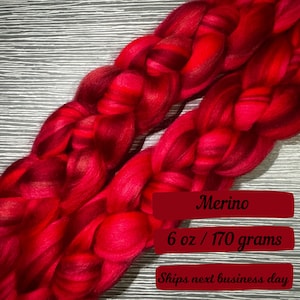 May include: Braided merino wool roving in vibrant shades of red. The fiber is 6 oz. (170 grams). The text "Merino" is displayed on a red banner.