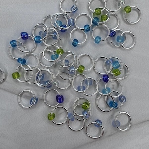 May include: A collection of silver ring stitch markers with blue, green, and clear beads. The markers are used for knitting and crocheting.