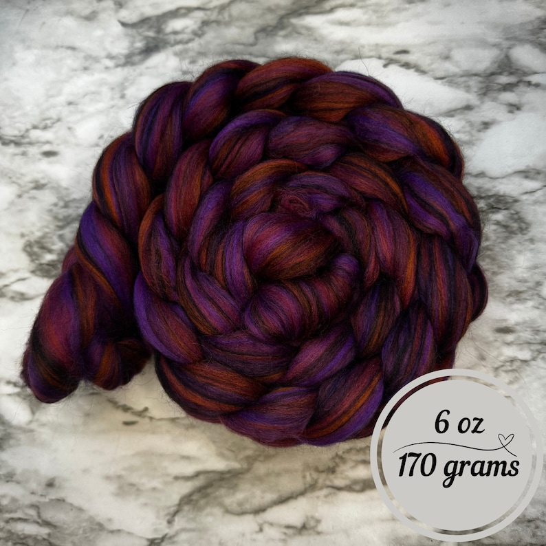 May include: A tightly coiled braid of colorful fiber in shades of deep purple, burgundy, and burnt orange. The image includes a circular graphic indicating the fiber weighs 6 oz (170 grams).