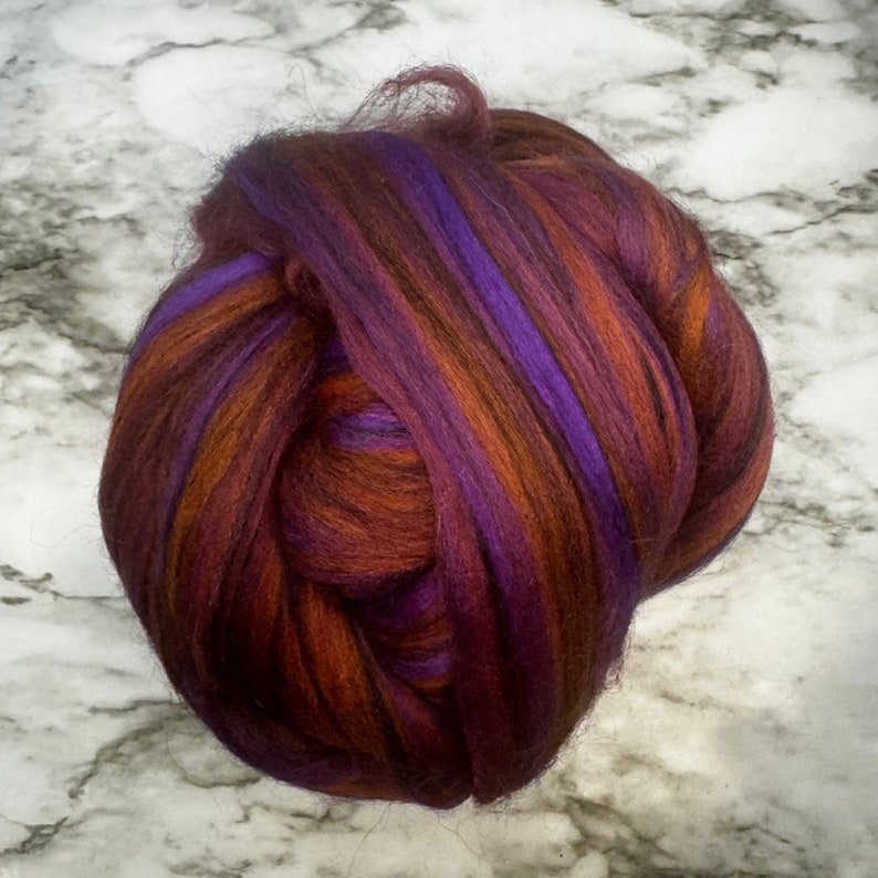 May include: A bundle of colorful fiber for crafting. The fiber is a mix of deep burgundy, vibrant purple, and warm orange hues. The fibers are tightly packed together, creating a textured appearance, ideal for various textile projects.