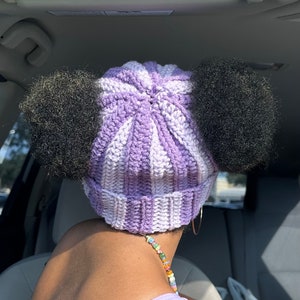 May include: A purple and white striped crochet beanie with two black fluffy pom-poms on top.