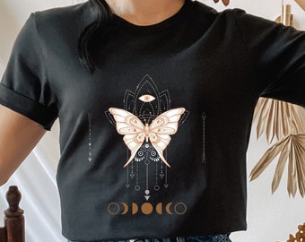 May include: Black t-shirt with a graphic of a butterfly with a moon and stars design. The shirt is worn by a person with dark hair and is paired with denim shorts.