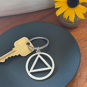 May include: A silver key chain with a key and a silver charm with a triangle inside a circle. The keychain is on a black plate.