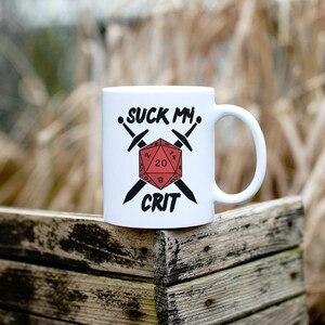 May include: White ceramic mug with the text "SUCK MY CRIT" in black, along with a red D20 die and crossed swords. The mug is a standard size and has a curved handle. The design is centered on the mug.