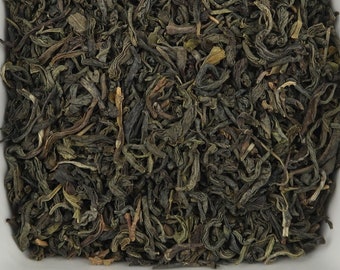 May include: A close-up of loose green tea leaves in a white ceramic bowl. The tea leaves are a dark green colour and have a slightly brown hue.