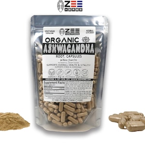 May include: A silver pouch containing organic Ashwagandha root capsules. The pouch has a label with the brand name "Zee Herbs" and the text "Organic Ashwagandha Root, Capsules" and "Supports Overall Health & Vitality". The pouch is sitting on a white surface. There is a pile of brown powder and a small pile of capsules to the left and right of the pouch.