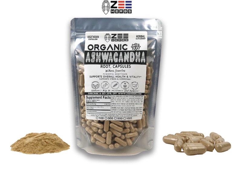 May include: A silver pouch containing organic Ashwagandha root capsules. The pouch has a label with the brand name "Zee Herbs" and the text "Organic Ashwagandha Root, Capsules" and "Supports Overall Health & Vitality". The pouch is sitting on a white surface. There is a pile of brown powder and a small pile of capsules to the left and right of the pouch.
