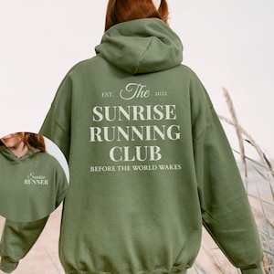 Puede incluir: Sudadera con capucha verde oliva con el texto "The Sunrise Running Club" impreso en la espalda. La sudadera con capucha también presenta el texto "Before the World Wakes" y "Est. 2022". Una imagen más pequeña muestra el texto "Sunrise Runner" en la parte delantera.