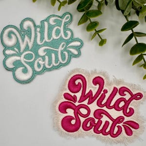 May include: Two embroidered patches with the text "Wild Soul" in different colors. The top patch is turquoise with white lettering and the bottom patch is white with pink lettering and a frayed edge.