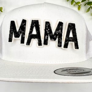 Op de afbeelding: Witte truckercap met een zwarte 'MAMA' patch van pailletten op de voorkant.