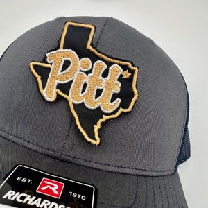 May include: A gray and black mesh trucker hat with a gold and white embroidered patch of the state of Texas with the word "Pitt" inside. The hat has a black mesh back and a black brim. The hat has a red and white label on the front that says "EST. Richards 1970".