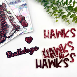 Op de afbeelding: Een set rode pailletten strijk-op patches, inclusief de woorden "Bulldogs" en "Hawks". De patches zijn verpakt in doorzichtige plastic zakjes.