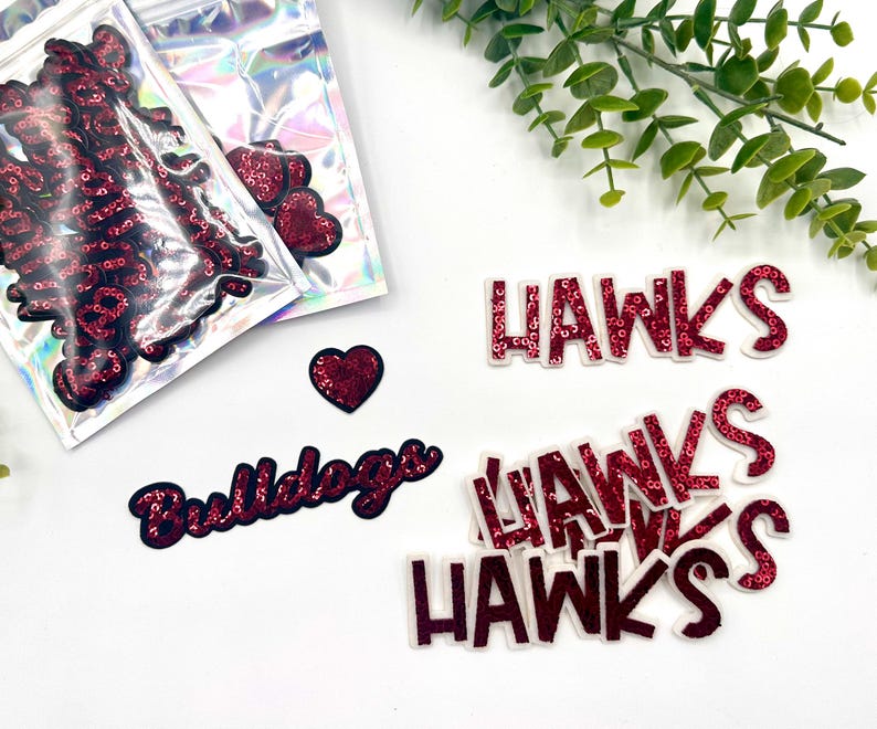 Op de afbeelding: Een set rode pailletten strijk-op patches, inclusief de woorden "Bulldogs" en "Hawks". De patches zijn verpakt in doorzichtige plastic zakjes.