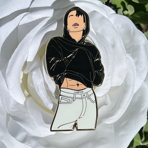 May include: Gold-colored enamel pin featuring a cartoon illustration of a person wearing a black hoodie and white jeans. The person has black hair and is wearing a silver earring. The text "Jungkook" is printed on the waistband of the jeans.