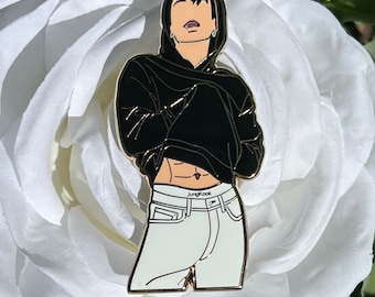 May include: Gold-colored enamel pin featuring a cartoon illustration of a person wearing a black hoodie and white jeans. The person has black hair and is wearing a silver earring. The text "Jungkook" is printed on the waistband of the jeans.