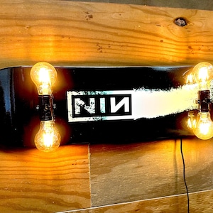 May include: A black skateboard deck wall light featuring the text NIN in a white rectangular box. The deck is illuminated by four glowing incandescent light bulbs mounted in pairs at each end, with a black power cord extending from the bottom.