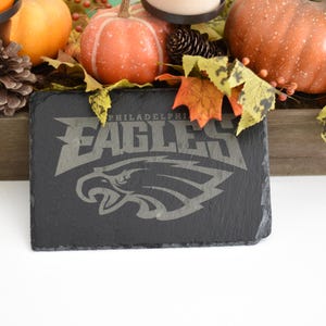 Puede incluir: Un posavasos de pizarra negra con el logotipo de los Philadelphia Eagles en gris. El posavasos forma parte de un arreglo temático otoñal con calabazas, hojas y piñas. El arreglo está sobre una superficie de madera.