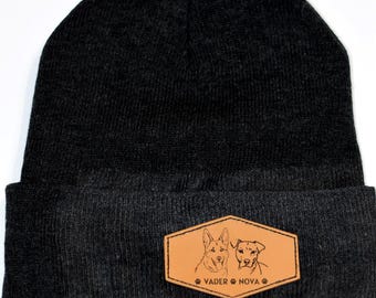 May include: Black knit beanie with a matching pom-pom on top. A brown leather patch on the front features line drawings of two dogs and the text "VADER NOVA".