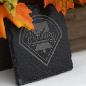 Puede incluir: Un posavasos de pizarra negra con el logotipo de los Phillies grabado en la superficie. El logotipo presenta la palabra "Phillies" en una fuente de escritura, con una Campana de la Libertad y un diseño de diamante de béisbol. El posavasos es cuadrado y tiene una superficie texturizada.